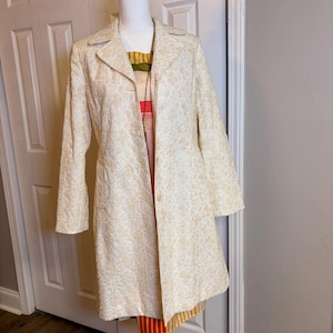 May include: A cream-colored long-sleeve coat featuring a textured floral pattern, a notched collar, and two visible buttons down the front. Underneath the coat, a multi-colored dress with horizontal stripes in yellow, green, and red is visible.