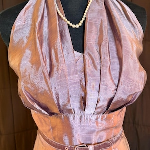 May include: A vintage-style, lavender-colored dress with a halter neckline and a fitted waist, cinched with a brown belt. A delicate pearl necklace adorns the neckline. The dress has a shimmering, textured fabric, suggesting a formal or special occasion garment.