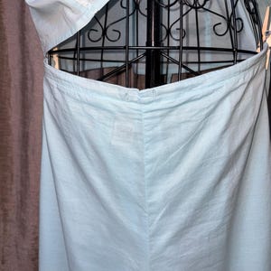 May include: Light blue garment, possibly a dress or top, displayed on a black metal dress form. The fabric appears to be lightweight, with visible seams and a button closure. The garment is partially open, revealing the dress form.