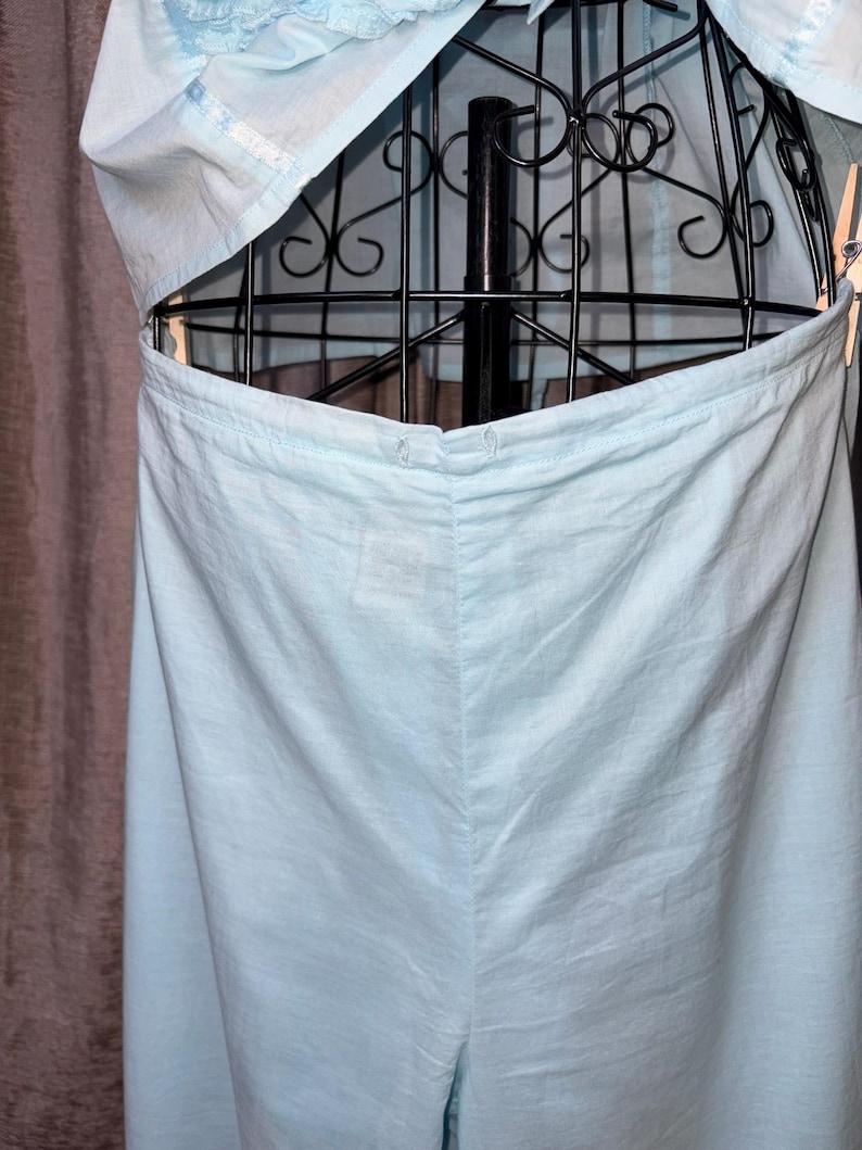 May include: Light blue garment, possibly a dress or top, displayed on a black metal dress form. The fabric appears to be lightweight, with visible seams and a button closure. The garment is partially open, revealing the dress form.