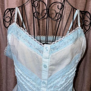 May include: A light blue and white camisole with lace trim and button details. The camisole is displayed on a black metal dress form. The garment features delicate lace along the neckline and straps, with a ruffled design.