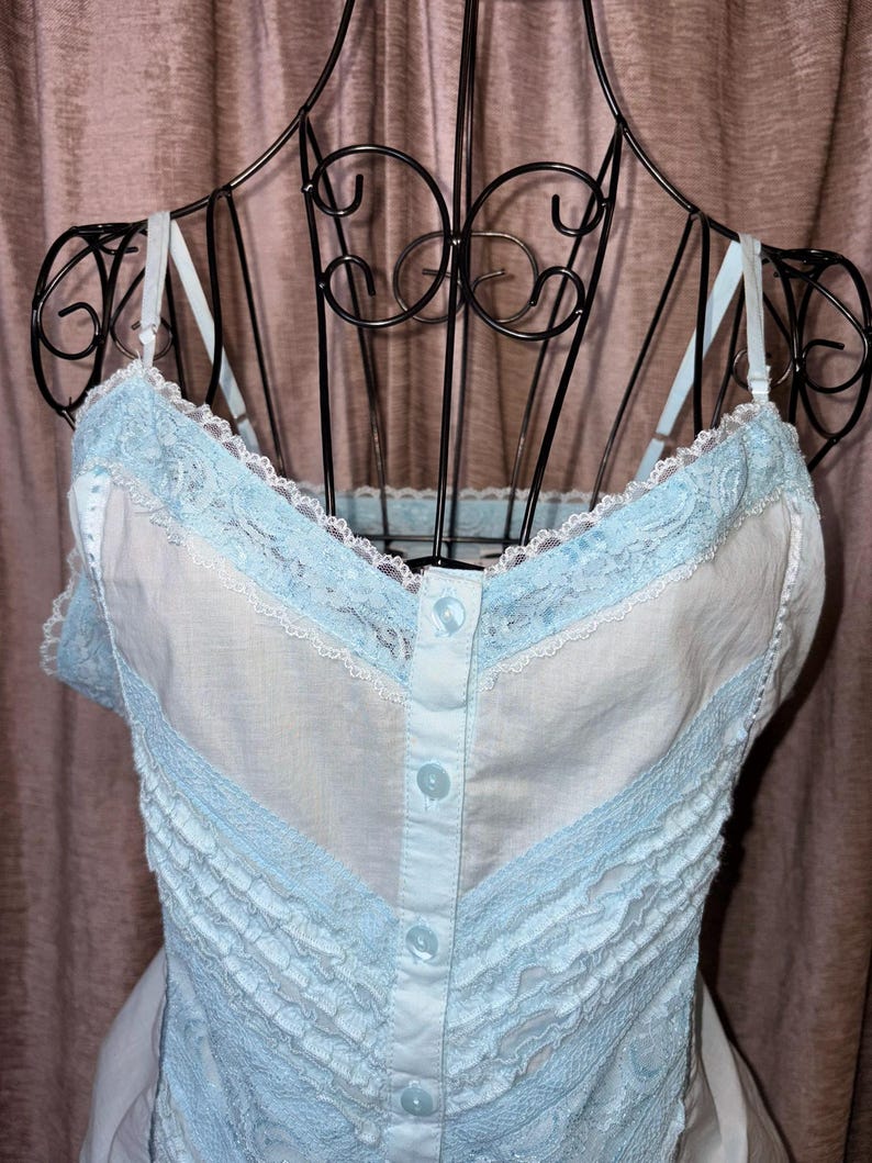 May include: A light blue and white camisole with lace trim and button details. The camisole is displayed on a black metal dress form. The garment features delicate lace along the neckline and straps, with a ruffled design.