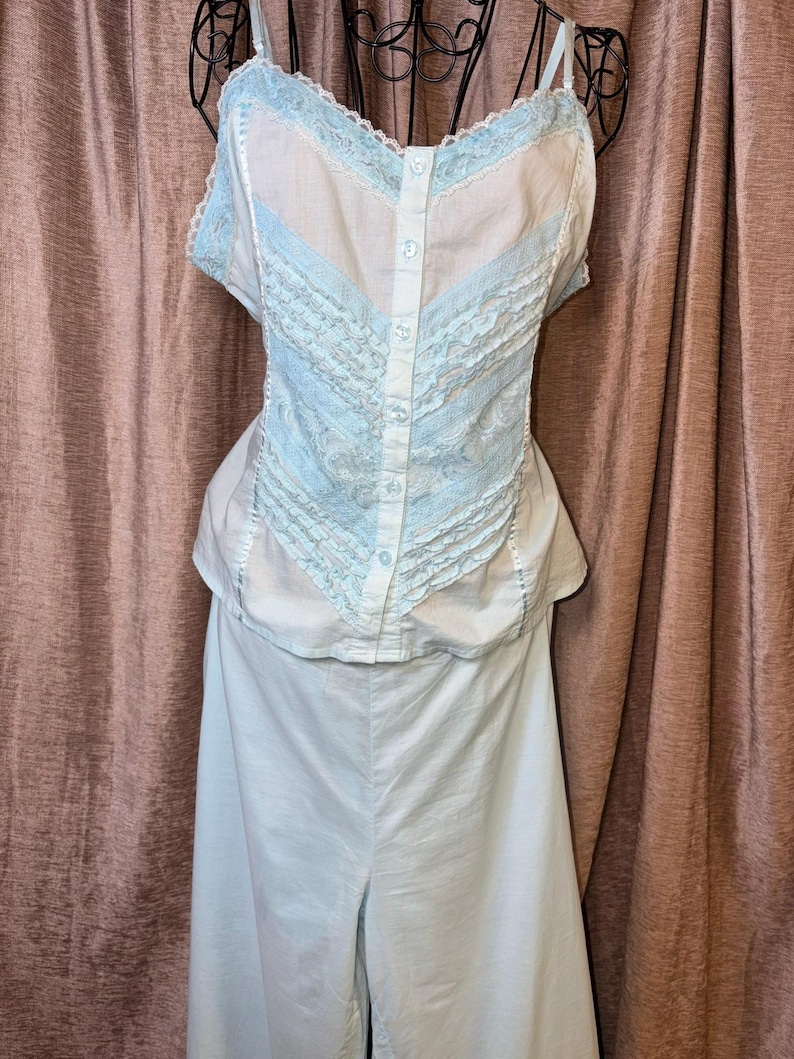 May include: A light blue two-piece lingerie set. The top has a button-down front with light blue lace trim and spaghetti straps. The matching bottoms are wide-legged pants. The set is displayed against a brown curtain.