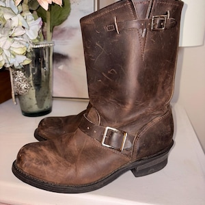 May include: Brown leather motorcycle boots with buckle details. The boots have a worn, distressed appearance, with visible scuffs and creases. The boots are mid-calf height and feature a rounded toe and a sturdy sole.