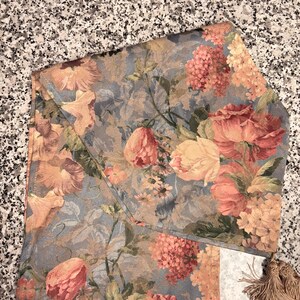 Puede incluir: Camino de mesa floral con un estampado de flores rosas, melocot&oacute;n y amarillas sobre un fondo azul gris&aacute;ceo. El camino de mesa tiene forma rectangular con borde doblado y flecos.