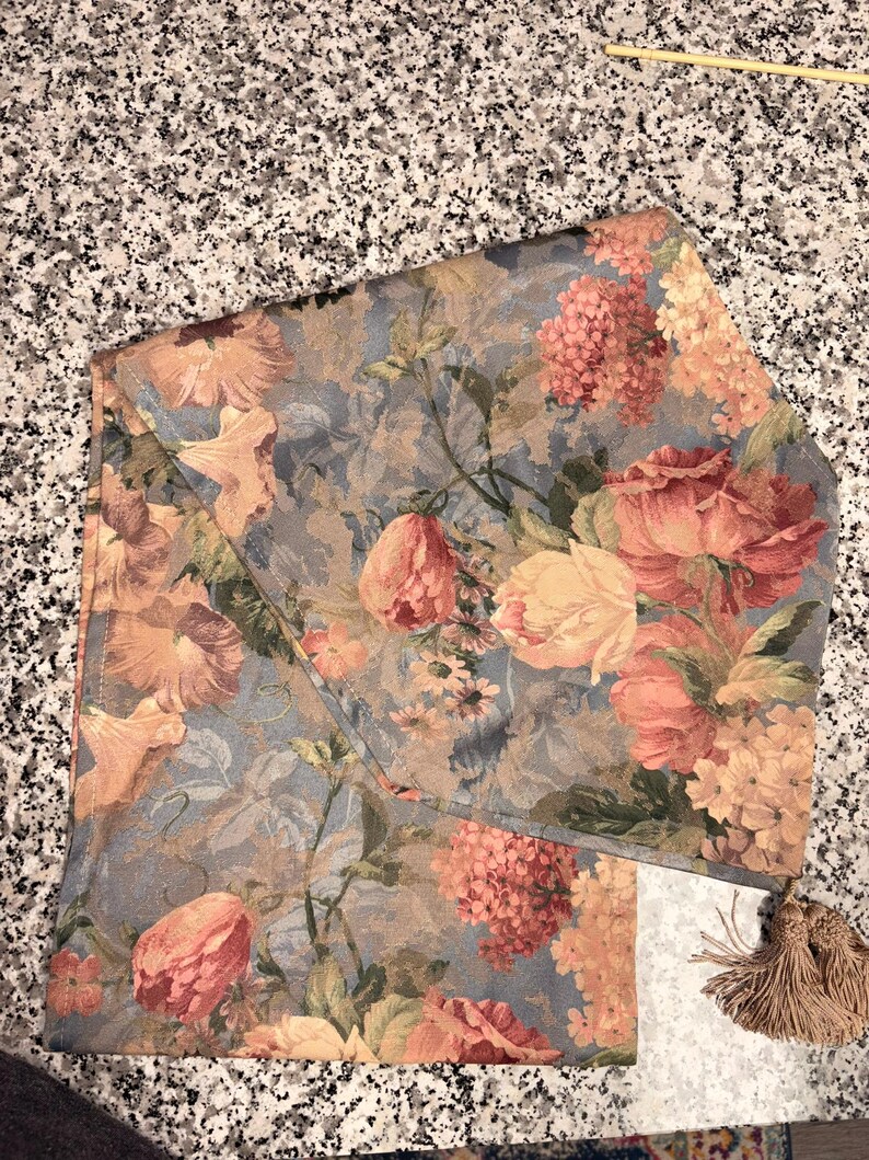Puede incluir: Camino de mesa floral con un estampado de flores rosas, melocot&oacute;n y amarillas sobre un fondo azul gris&aacute;ceo. El camino de mesa tiene forma rectangular con borde doblado y flecos.