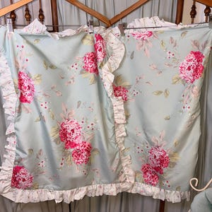 May include: Two light blue pillowcases with a floral pattern of pink hydrangeas and green leaves. The pillowcases have a ruffled edge in white with a floral print. The pillowcases are hanging on wooden hangers.