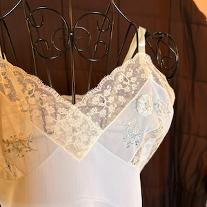 May include: A light blue vintage slip with delicate ivory lace trim and embroidered floral details. The slip is displayed on a black metal dress form, showcasing its elegant design and soft fabric.