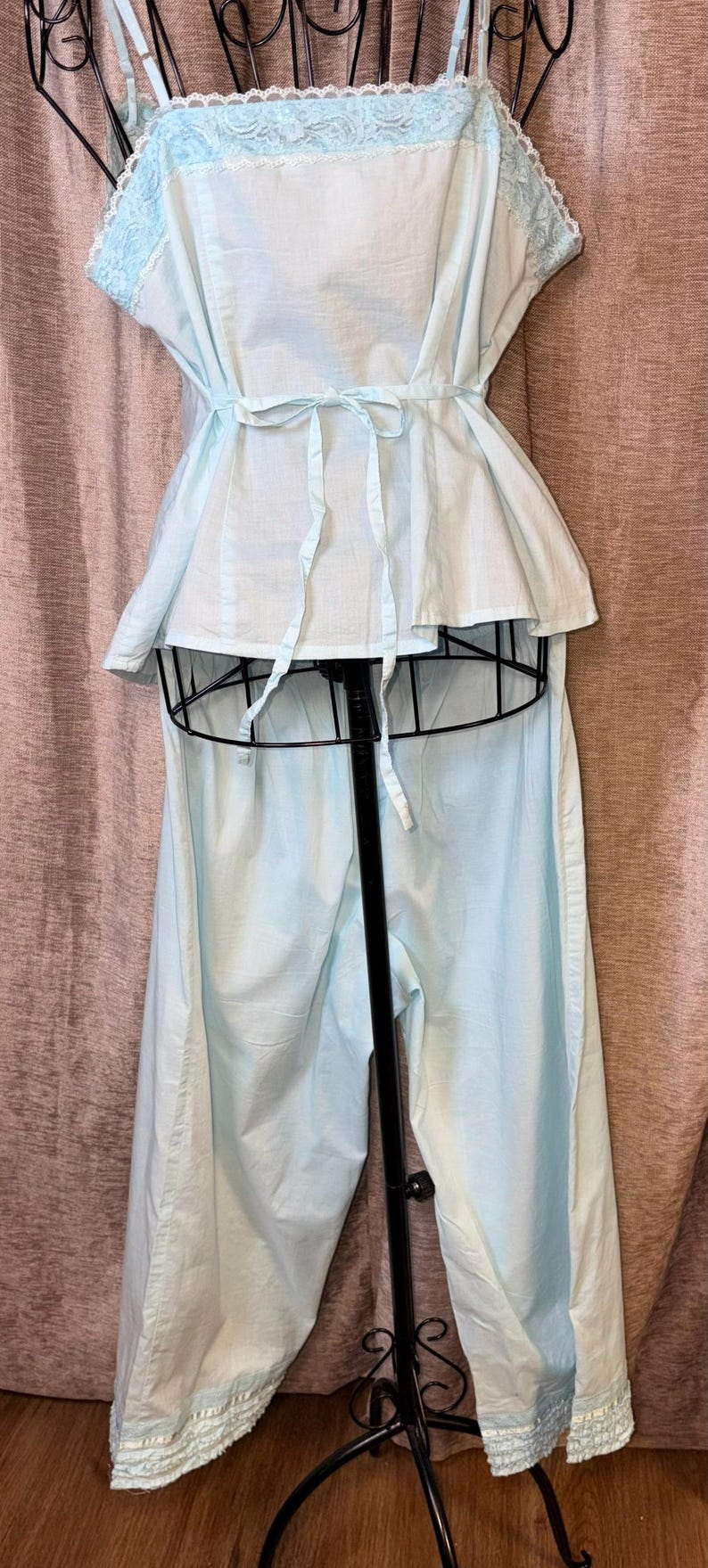 May include: A light blue vintage pajama set. The top has a peplum waist and tie closure. The camisole top has lace trim. The pajama pants are wide-legged. The set is displayed on a black mannequin.