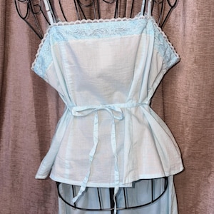 May include: Light blue vintage-style pajama set. The top features a square neckline with delicate lace trim and adjustable straps. A matching tie belt cinches the waist. The pajama bottoms are visible below the top. The set is displayed on a black mannequin.
