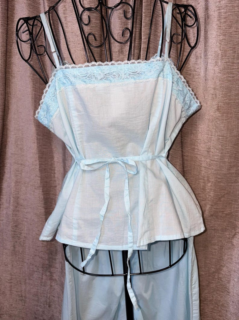May include: Light blue vintage-style pajama set. The top features a square neckline with delicate lace trim and adjustable straps. A matching tie belt cinches the waist. The pajama bottoms are visible below the top. The set is displayed on a black mannequin.