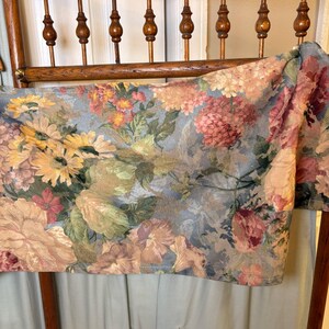 Puede incluir: Una tela estampada con flores drapeada sobre el respaldo de una silla de madera. La tela presenta una variedad de flores en tonos rosa, amarillo y melocot&oacute;n sobre un fondo azul gris&aacute;ceo. El respaldo de la silla es de madera.