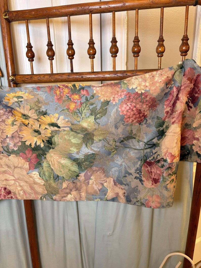 Puede incluir: Una tela estampada con flores drapeada sobre el respaldo de una silla de madera. La tela presenta una variedad de flores en tonos rosa, amarillo y melocot&oacute;n sobre un fondo azul gris&aacute;ceo. El respaldo de la silla es de madera.