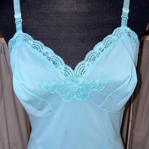 May include: A light blue slip dress featuring a V-neckline adorned with matching lace trim and a central lace appliqué. The garment includes adjustable shoulder straps with white plastic sliders, providing a secure fit for the bodice area.
