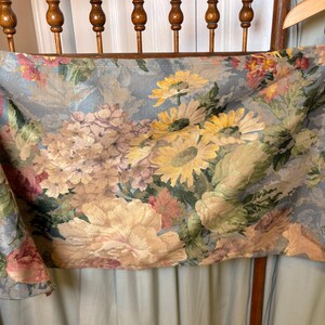 Puede incluir: Una tela floral con un fondo azul claro, con grandes girasoles amarillos, peon&iacute;as rosas y melocot&oacute;n, y otras flores coloridas. La tela est&aacute; drapeada sobre una silla de madera.