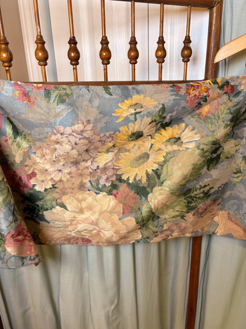 Puede incluir: Una tela floral con un fondo azul claro, con grandes girasoles amarillos, peon&iacute;as rosas y melocot&oacute;n, y otras flores coloridas. La tela est&aacute; drapeada sobre una silla de madera.