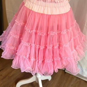 May include: A pink tulle skirt with multiple layers of ruffles. The skirt has a wide, light pink waistband and is displayed on a white mannequin. The top portion of the skirt is a floral pattern.
