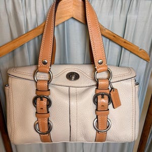 May include: A cream-colored leather handbag featuring tan leather straps and trim. The bag includes silver-toned metal hardware, two vertical buckled straps on the front, a central turn-lock closure, and a small leather luggage tag attached to a ring.