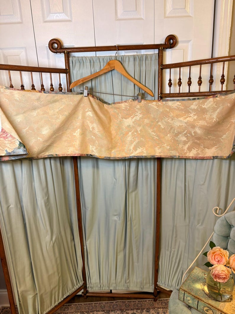 Puede incluir: Un biombo de madera vintage con un fondo de tela azul claro. Una tela estampada est&aacute; drapeada sobre la parte superior, sujeta por una percha de madera. El biombo tiene un dise&ntilde;o decorativo y ornamentado.