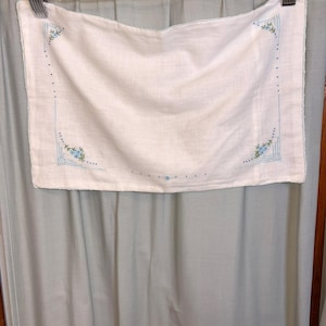 May include: A rectangular white fabric piece featuring delicate blue floral embroidery at the corners and along the edges. The design includes small blue flowers, green leaves, and thin blue stitched lines forming a border frame on the cloth.
