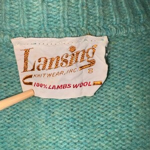 Może przedstawiać: Zbliżenie turkusowego swetra. Etykieta głosi "Lansing Knitwear, Inc." i "100% Lambs Wool". Sweter ma prążkowany dekolt i wzorzysty wz&oacute;r dzianiny. Drewniany wskaźnik dotyka etykiety.