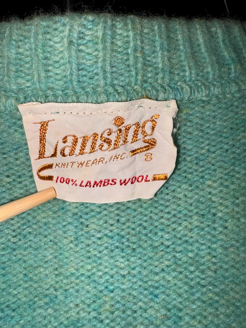 Może przedstawiać: Zbliżenie turkusowego swetra. Etykieta głosi "Lansing Knitwear, Inc." i "100% Lambs Wool". Sweter ma prążkowany dekolt i wzorzysty wz&oacute;r dzianiny. Drewniany wskaźnik dotyka etykiety.