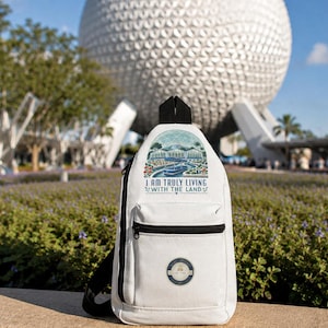 May include: A white sling backpack featuring a black zipper and a graphic print of a greenhouse structure with the text I AM TRULY LIVING WITH THE LAND. The bag has a small circular patch on the front pocket and a black strap attached to the top.