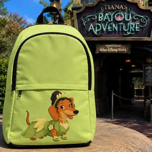 May include: A lime green backpack featuring a graphic of a brown dachshund wearing a green dress and a tiara. The backpack has a large main compartment and a smaller front pocket, both secured with black zips and black trim along the edges.