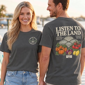 May include: A charcoal gray short-sleeve t-shirt featuring a small circular logo on the front chest and a large graphic on the back. The back design displays domed greenhouses, pumpkins, tomatoes, and lemons with the text LISTEN TO THE LAND.