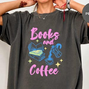 May include: A charcoal gray oversized t-shirt featuring the pink text Books and Coffee. The graphic includes a stack of blue books, a blue moka pot pouring coffee into a cup, and small yellow stars. The shirt has a crew neckline and short sleeves.