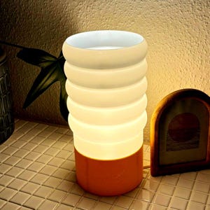 Könnte beinhalten: Eine zylindrische Tischlampe mit einem gestapelten, ringförmigen Design. Der Sockel ist warmorange, geht in ein leuchtendes, cremefarbenes Licht über. Die Lampe steht auf einer gefliesten Oberfläche, mit einem gerahmten Bild in der Nähe.