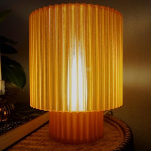 Könnte beinhalten: Eine zylindrische Tischlampe in Bernstein mit geripptem Schirm und Sockel. Die Lampe ist beleuchtet und strahlt ein warmes Licht aus. Die Lampe steht auf einer gewebten Oberfläche.