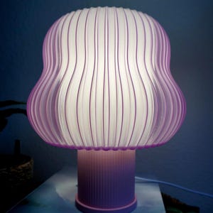 Può includere: Lampada da tavolo decorativa con paralume a forma di fungo. La lampada presenta un paralume viola traslucido e a coste con linee verticali e una base viola abbinata. La lampada è accesa, emettendo un bagliore morbido e caldo.