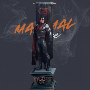 May include: A detailed statue of a superhero in a black and red suit, standing on a pedestal. The statue has a red cape and a banner with a symbol. The word "MANUAL" is written in orange.