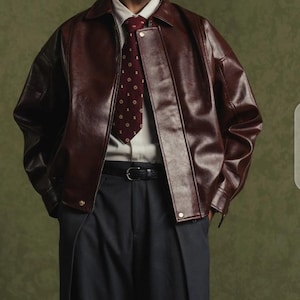 Puede incluir: Chaqueta de cuero burdeos con cuello, usada sobre una camisa blanca y una corbata estampada. La chaqueta tiene cierre de cremallera y se combina con pantalones grises oscuros y un cinturón negro. El conjunto se encuentra sobre un fondo verde.