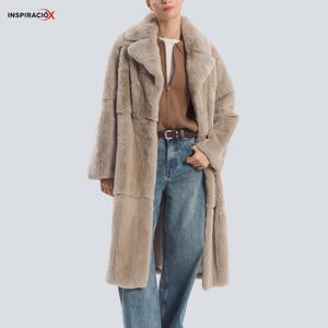 May include: A long, beige faux fur coat with a wide collar and large lapels. The coat is worn over a brown sweater and blue jeans. The coat has a patchwork design and a belt at the waist. The word "INSPIRACIÓX" is in the top left corner.