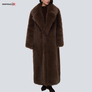 May include: A long, brown faux fur coat with a wide lapel collar. The coat has long sleeves and a straight cut. The coat is a solid colour and appears to be made of a soft, plush material. The word "INSPIRACIÓX" is in the top left corner.