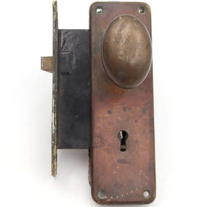May include: An antique door lock with a tarnished brass finish. The lock features an oval-shaped doorknob and a keyhole. The rectangular plate shows signs of age and wear, with a dark, aged appearance. The lock is mounted on a white background.