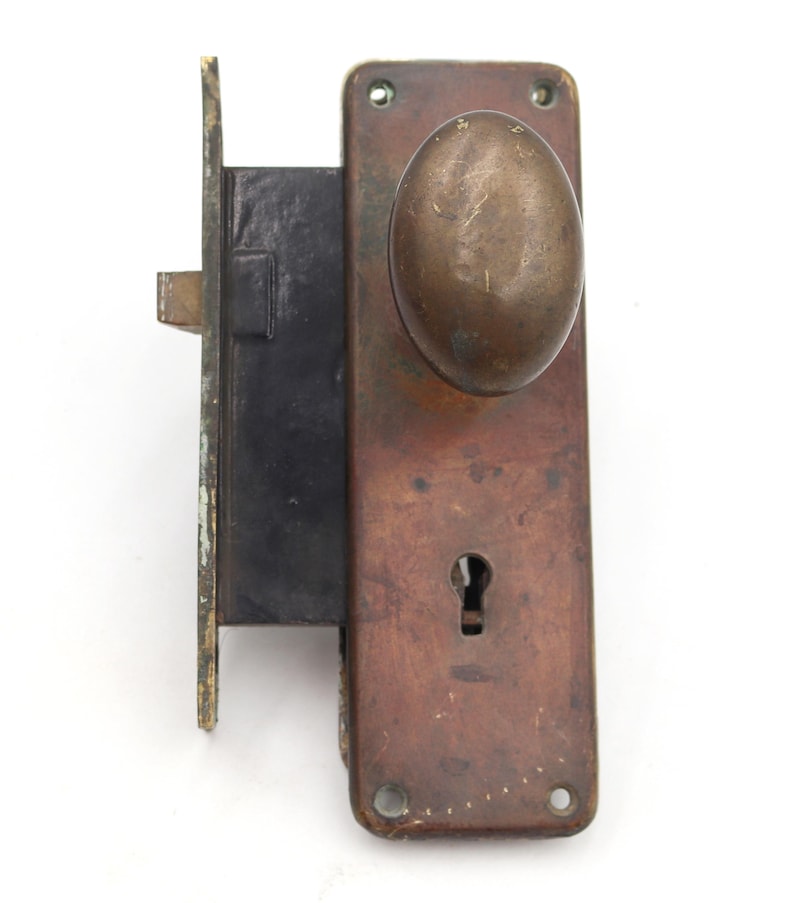 May include: An antique door lock with a tarnished brass finish. The lock features an oval-shaped doorknob and a keyhole. The rectangular plate shows signs of age and wear, with a dark, aged appearance. The lock is mounted on a white background.