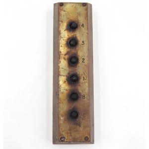 May include: An antique elevator control panel with a rectangular brass face. It features six black buttons labelled with numbers 1-4, and letters B and C. The panel shows signs of age and wear.