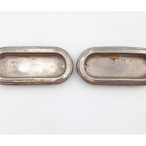 May include: Two antique, oval-shaped metal drawer pulls with a tarnished, silver finish. Each pull has a recessed center and two small holes for mounting. The pulls are set against a white background.