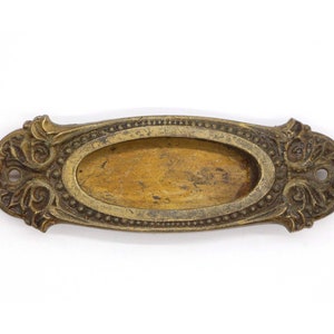 Puede incluir: Una placa de puerta ovalada de latón con un diseño ornamentado. La placa tiene un borde elevado y una superficie lisa y pulida.