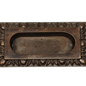 Könnte beinhalten: Ein rechteckiger bronzener Schrankgriff mit einem verzierten Design. Der Griff hat eine vertiefte ovale Öffnung für einen Fingergriff.
