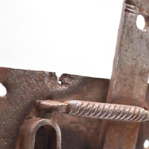 May include: Close-up of a weathered, rusty metal latch. The latch features a rectangular plate with a curved handle and a sliding bar. The metal has visible wear and tear, with a textured surface and signs of age.