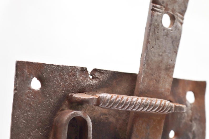 May include: Close-up of a weathered, rusty metal latch. The latch features a rectangular plate with a curved handle and a sliding bar. The metal has visible wear and tear, with a textured surface and signs of age.