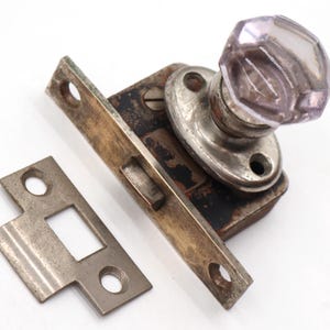 May include: Antique door hardware set featuring a faceted, lavender glass doorknob. The set includes a latch, strike plate, and mounting hardware, all with a vintage, aged appearance. The metal components show signs of wear and patina.