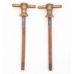 May include: Two identical metal T-handle bolts featuring a brass-toned top handle with a flared neck and a long, threaded steel shaft. The threaded sections exhibit a rusted, textured brown finish, while the handles have a smooth, metallic sheen.