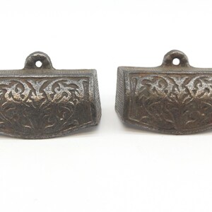 May include: Two antique cast iron drawer pulls with a decorative floral design. The pulls are a dark grey colour and have a slightly aged appearance.