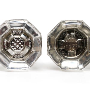 Pode incluir: Dois puxadores de porta de vidro transparente, octogonais, com base de metal prateado. Os puxadores têm um design circular central com pequenos pontos em relevo. O vidro tem um aspeto facetado, refletindo a luz.