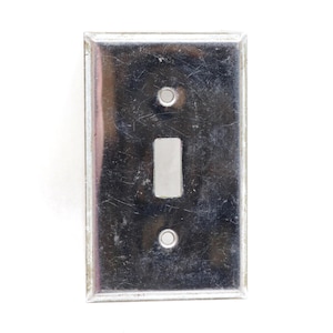 May include: A rectangular, metallic silver light switch wall plate with a reflective, polished finish. The plate features a central vertical rectangular cutout for a toggle switch and two circular screw holes positioned above and below the center.